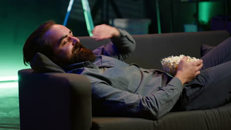 Amused man lounged on couch laughing at funny comedy movie while eating popcorn