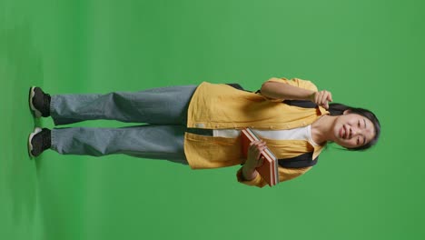 full body of asian woman student with a backpack and some books looking at the camera and shouting at someone while standing in the green screen background studio