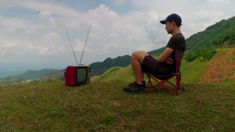 Lustiges-Video-Eines-Mannes,-Der-In-Einem-Kleinen-Stuhl-Mit-Bergkulisse-Sitzt-Und-Einen-Alten-Fernseher-Anschaut