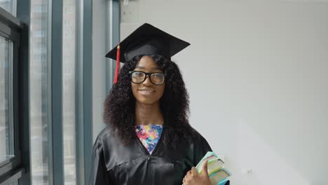 joven graduada universitaria afroamericana de pie con libros de texto en las manos cerca de una gran ventana moderna con marcos negros. estudiante de sexo femenino con una túnica negra, sombrero de maestro cuadrado y gafas