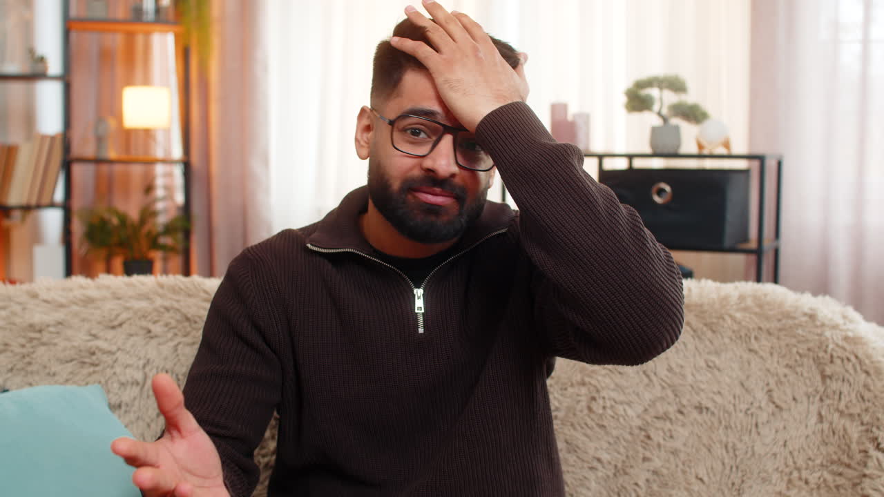 Un homme indien sur le canapé à la maison couvre le visage avec sa main dans la frustration réagissant avec un geste de facepalm