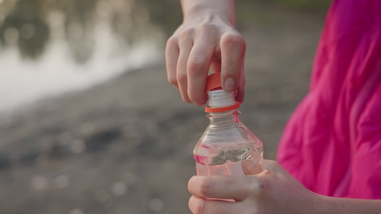 Retkeilijä avaa vesipulloa järven rannalla, kädet kiertämässä korkkia irti pienestä muovipullosta, vaaleanpunainen takki ja vihreä hiha näkyvissä, pikkukivinen ranta ja tyyni järvi taustalla, hienovarainen huurtuminen.