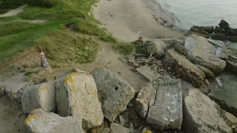 Aerial-establishing-view-of-beautiful-young-romantic-caucasian-girl-in-a-long-dress-on-the-white-sand-beach-with-old-ruins,-sunny-summer-evening,-sunset,-golden-hour,-wide-drone-orbit-shot