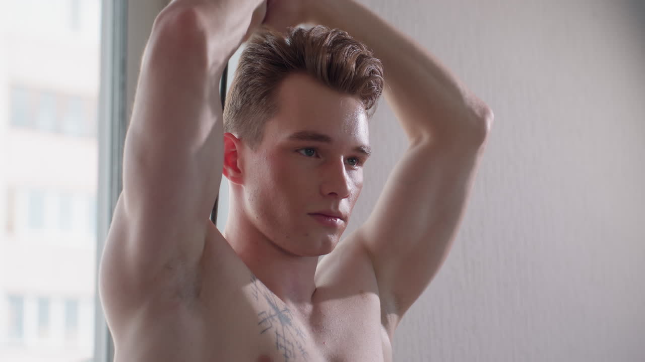 close up of gym lover standing near window pulling resistance band overhead with focused expression, sunlight gently filtering through glass highlighting defined chest muscles and tattooed skin