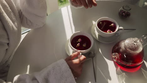 man adds rose buds into woman tea at table closeup. careful husband prepares romantic breakfast for wife at home in morning. domestic date atmosphere