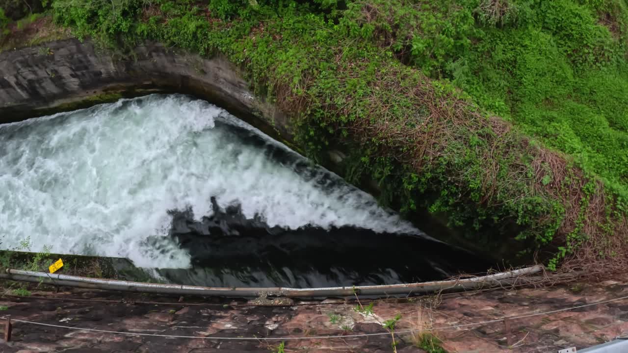concrete spillway where fast-moving Talvera water surges over a curved lip, churning white foam against mossy stone and verdant embankment, static camera looking down