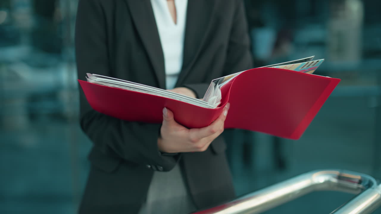 Partial view of lady in black blazer examining red folder outdoors with urban residential building in background, focus on hands and document, reflecting professional work setting