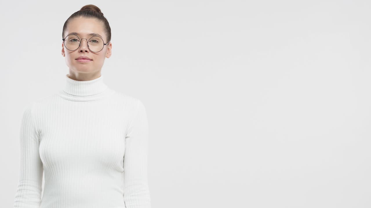 コピースペースのある灰色の背景に、丸い眼鏡をかけた若くておかしな少女が、右を指差し、驚きと幸せそうな表情で立っている。