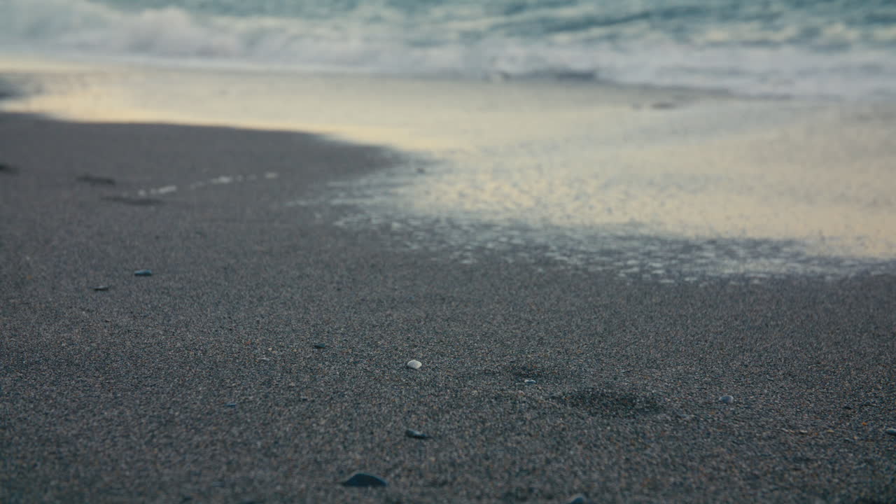 Close-up of a gentle wave washing away footprints on the beach, symbolizing impermanence and calm, captured in soft evening light