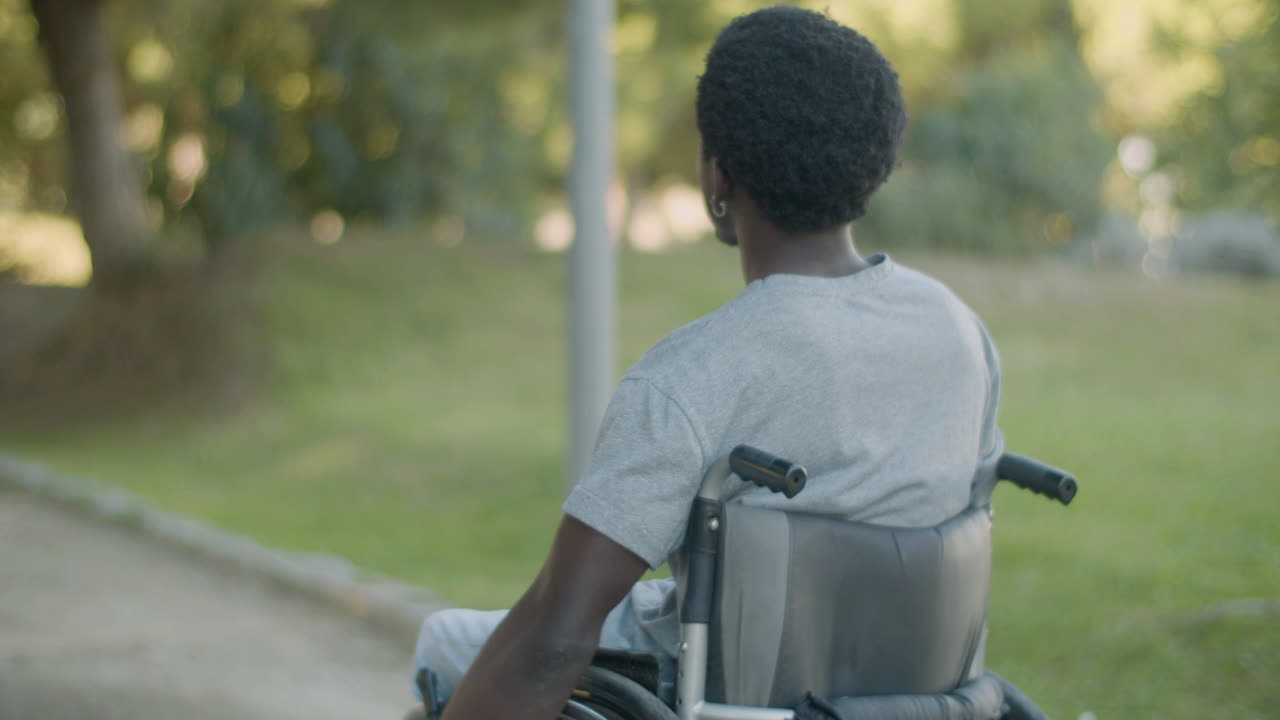 Back View Of Young Black Man Riding His Wheelchair In Park On Summer Day