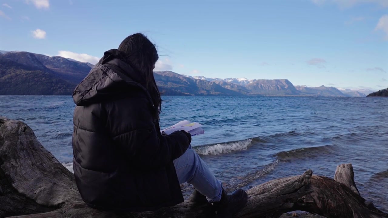 Woman reads beside Lake Traful in Villa Traful, Neuquén, Argentina, surrounded by crystal-clear water, driftwood, snow-capped Andes and tranquil Patagonian wilderness, static lakeside shot.