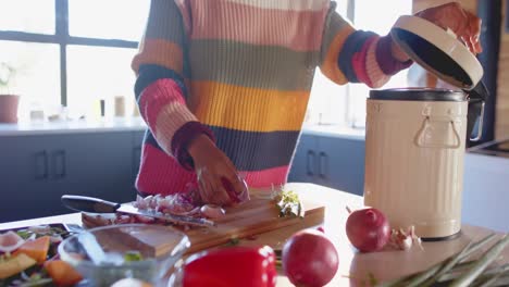 Mid-section-of-african-american-woman-composting-vegetable-waste-in-sunny-kitchen,-in-slow-motion