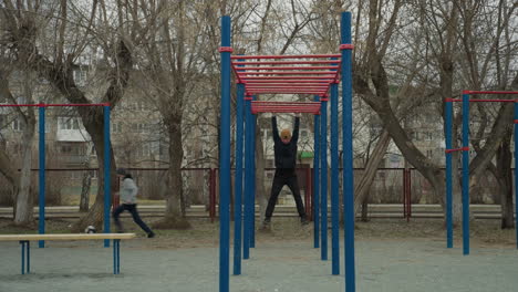a coach jumps up to grab a red iron bar and begins swinging in an outdoor exercise area, in the background, a residential building, passing cars, and someone playing football can be seen