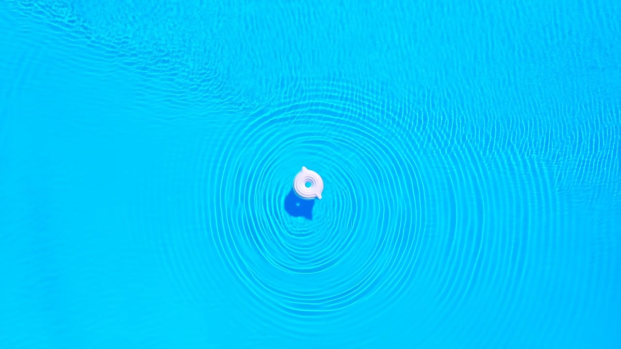 Pemandangan tampak atas cincin tiup putih kecil mengambang di kolam biru kehijauan cerah, menciptakan riak konsentris di permukaan air yang tenang sementara bayangan lembut dan pantulan jernih menciptakan kontras warna musim panas yang bersih