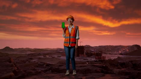 cuerpo lleno de ingeniera asiática con casco de seguridad sonriendo y mostrando pantalla verde teléfono inteligente a la cámara en el sitio de construcción, maquinaria de minería
