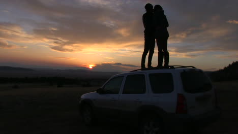 a silhouette of a couple standing on top of a suv kissing at sunset