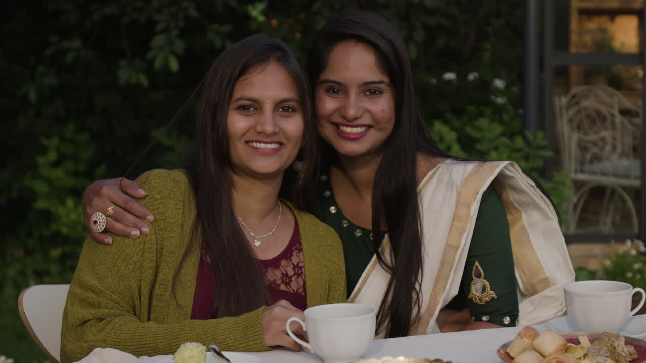 चाय और नाश्ते के साथ एक बगीचे में मिलन समारोह का आनंद लेती दो मुस्कुराती हुई भारतीय महिलाएँ।