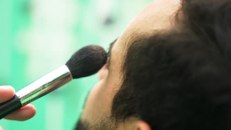 maquillaje de ojos hombre aplicando con un pincel un polvo de carnaval brillante en el rostro masculino con barba y fondo verde