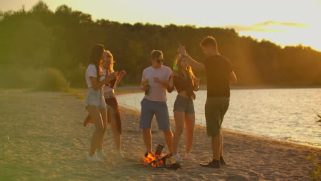 Die-Studenten-Tanzen-In-Shorts-Und-T-Shirts-Mit-Bier-Um-Das-Lagerfeuer-Am-Sandstrand.-Sie-Genießen-Den-Sommerabend-Am-Flussufer-Auf-Der-Open-Air-Party.