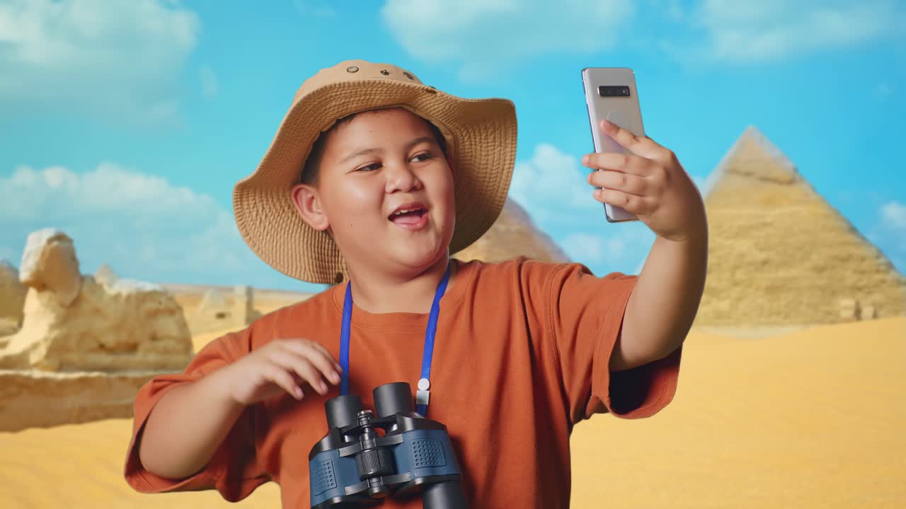 Asian Boy With A Hat And Binoculars Waving Hand Having Video Call On Smartphone While Traveling In Giza Pyramid. Boy Researcher Examines Something, Travel Adventure, Close Up