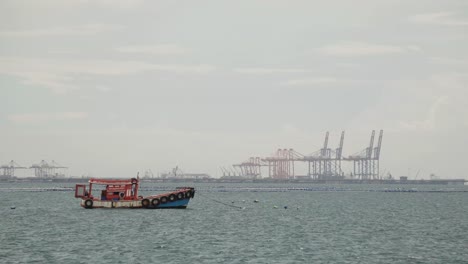 a moored thai fishing boat in the middle a bay, in the background the cranes of the sri rascha deep sea port, thailand