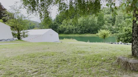 Grandes-Tiendas-De-Campaña-Blancas-Sobre-Un-Césped-Con-Algunos-árboles-Y-Un-Lago-Al-Fondo-Con-Bosque