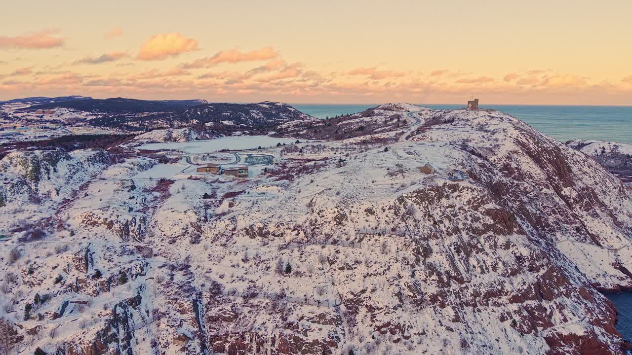 Flybilder av en snødekt kystklippe og åssideområde med pastellfarget himmel