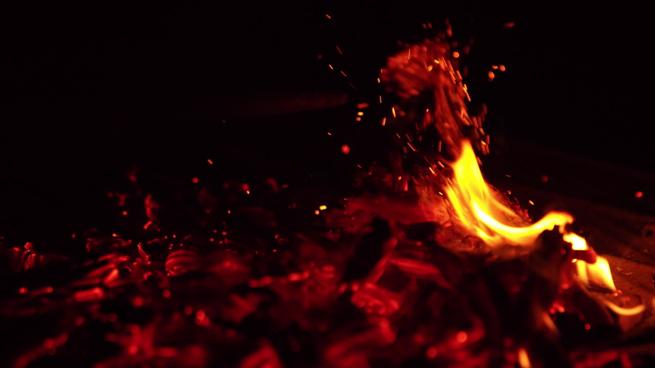 The campfire has burnt out and little flame is still on. Embers are moved with stick and beautiful sparks rise up. Close up. Black backdrop.
