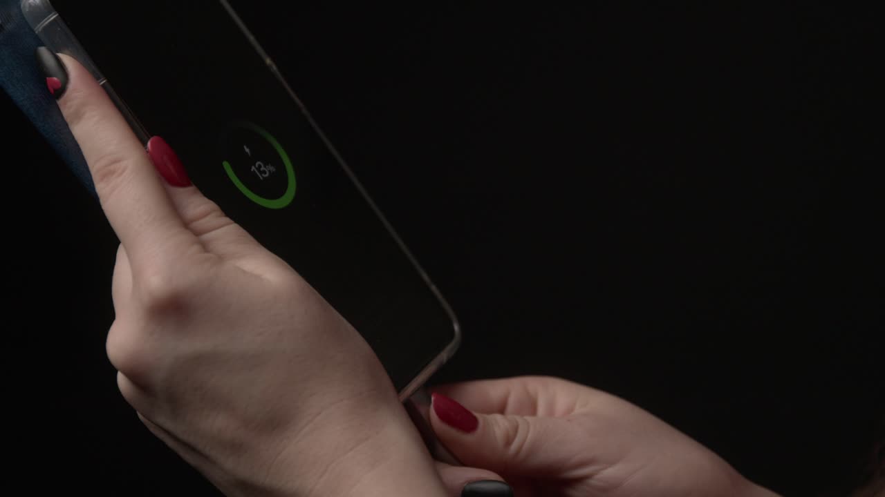 Detailed view of a smartphone being charged at 13%. Hands with red nails hold the device against a black background, creating a focused and tech-centric atmosphere