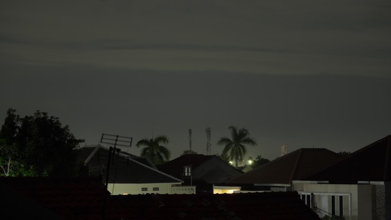 Vista noturna de casas, palmeiras, antenas e torres sob um céu escuro.