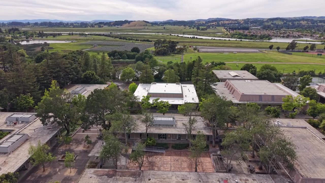 Veduta aerea che sorvola gli edifici e i terreni del Napa Valley College in California, mostrando il lussureggiante paesaggio verde circostante, il fiume e le dolci colline sotto un cielo parzialmente nuvoloso