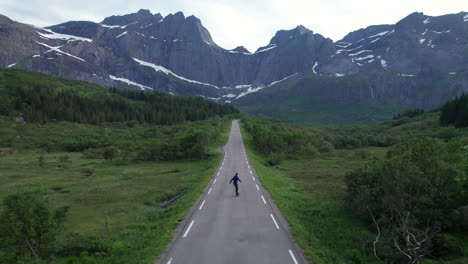 Aerial-Forwarding-shot-of-a-young-man-skating-alone-on-a-scenic-road-to-Nusfjord-in-Lofoten,-Norway