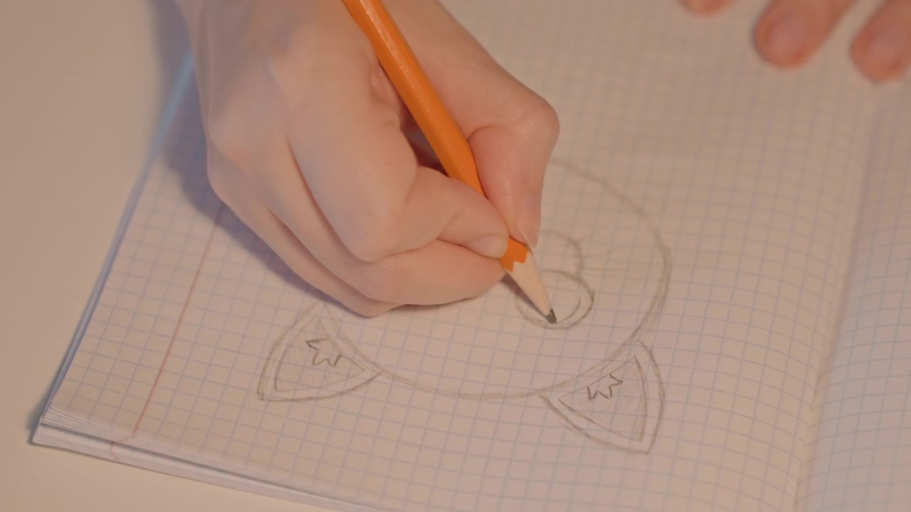 La mano sinistra abbozza un volto di animale da cartone animato con orecchie a punta. Il disegno procede costantemente mentre la matita arancione traccia le linee. La vista ravvicinata si concentra esclusivamente sulle mani e sulle pagine del quaderno