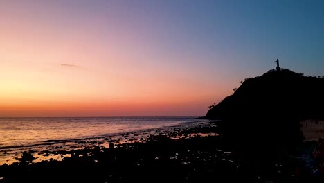 Silhouetted-Cristo-Rei-statue-of-Jesus-Christ-with-beautiful-pink-and-orange-sunset-over-peaceful-ocean-in-capital-Dili,-Timor-Leste,-Southeast-Asia