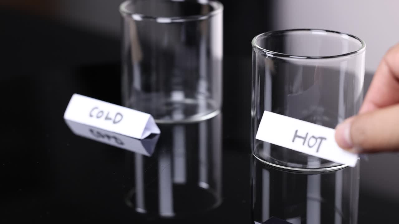 Hand places 'hot' and 'cold' paper labels on empty glass cups in a controlled environment