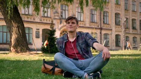 young handsome man is sitting on lawn in park, listening music with earphones, relax concept, building on background