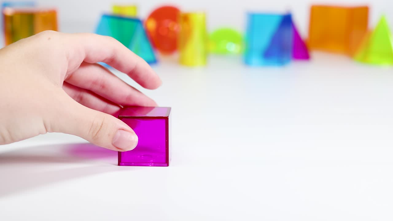 A hand manipulates a pink cube among colorful geometric shapes on a white surface, showcasing educational interaction