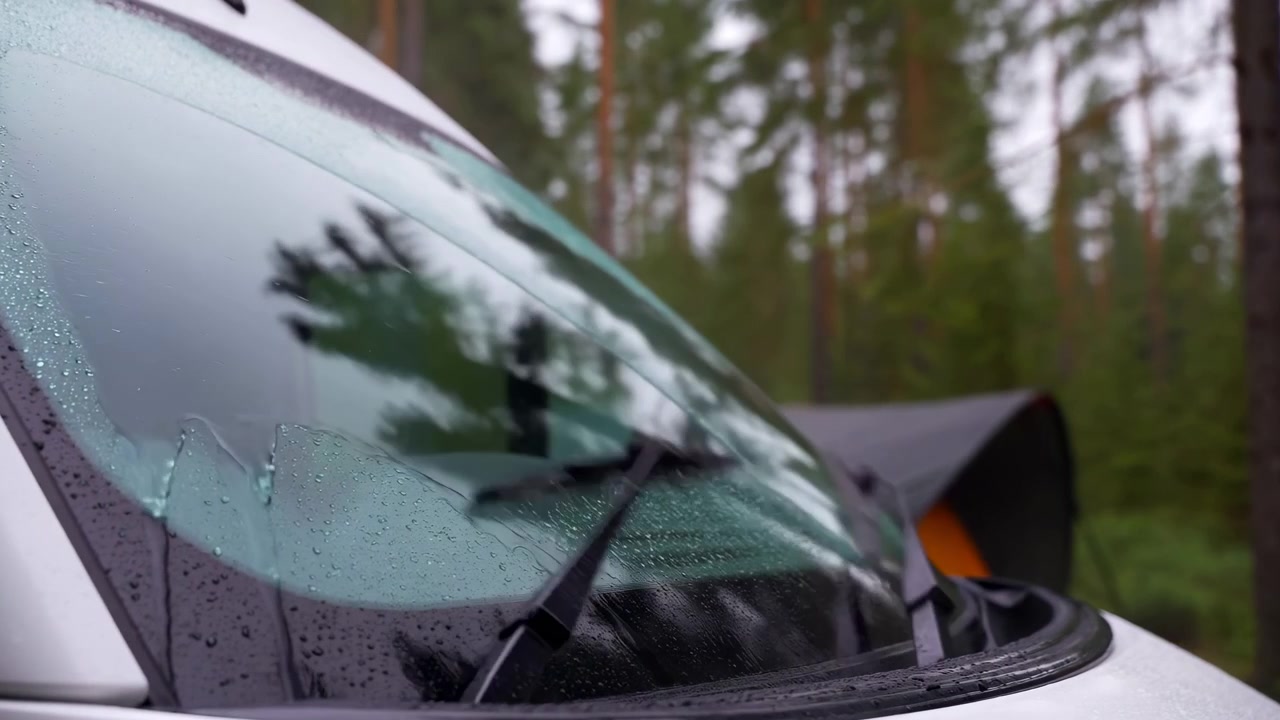 Una vista ravvicinata mostra i tergicristalli che spazzano via le goccioline di pioggia sul parabrezza mentre lo scenario della foresta verde rimane leggermente sfocato dietro il vetro, catturando il movimento del tempo umido e le superfici riflettenti.