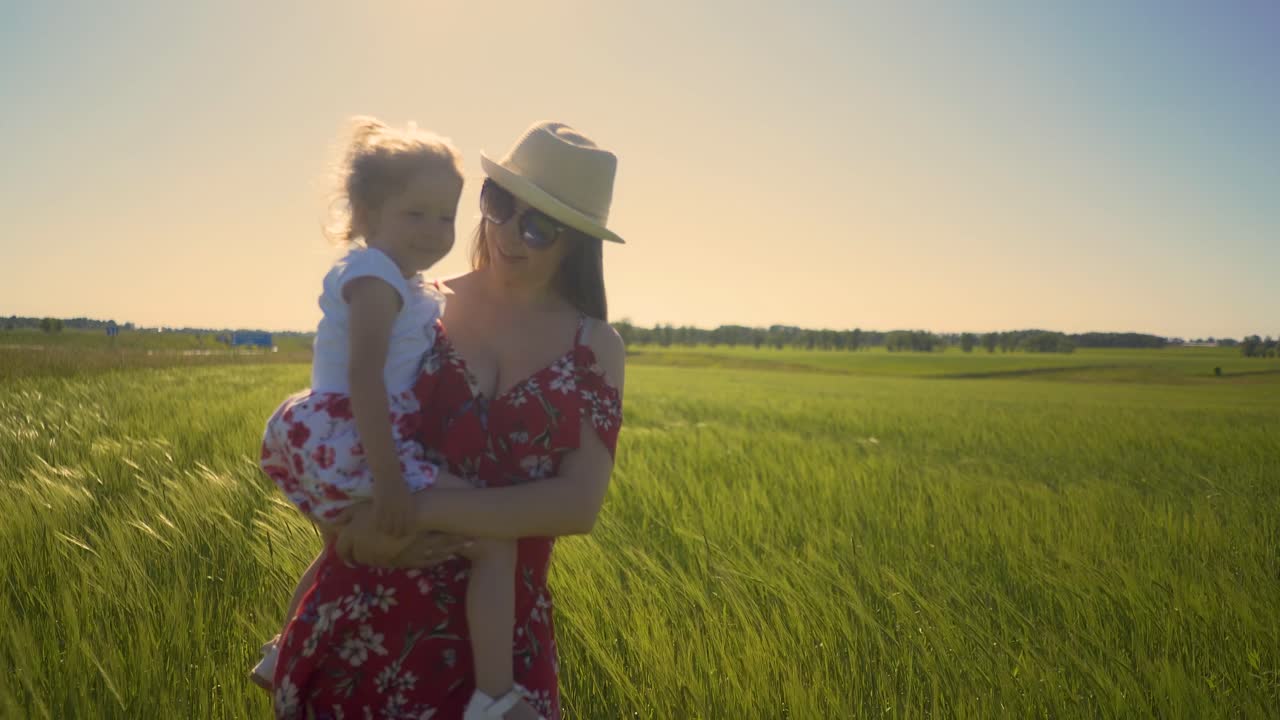 Verfolgende Nahaufnahme: Mutter im Sommerkleid und mit Sonnenbrille geht über ein weites Feld und trägt ihre kleine Tochter auf dem Arm.