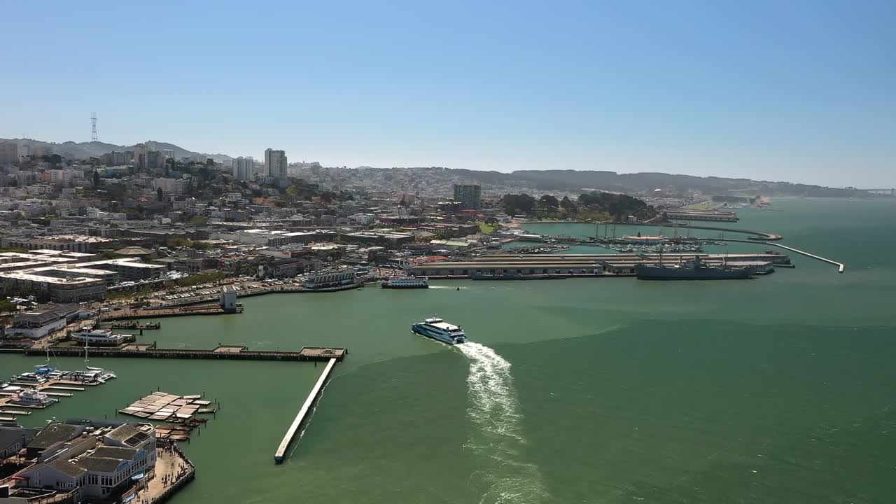 Aerial drone view showing ferry boat sailing across green waters of San Francisco Bay with city skyline, waterfront piers, and sunny clear sky in California USA