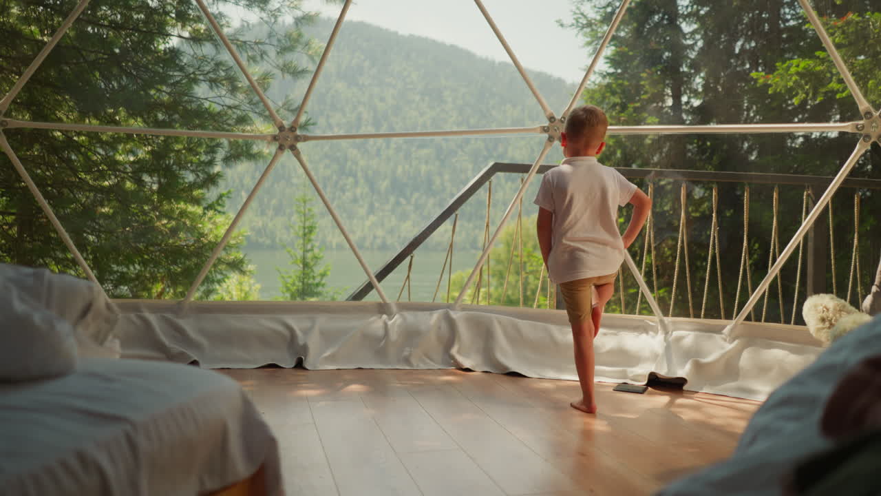Child exercises by panoramic window at glamping. Little boy does yoga standing in clear camp cabin. Cute kid sports activity on family vacation