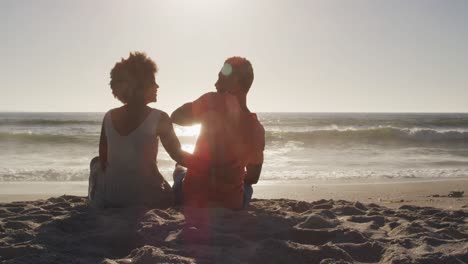 una pareja afroamericana sonriente abrazándose y sentada en una playa soleada