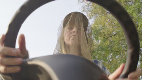 Lady-with-mosquito-head-net-on-tractor-mower-pestered-by-mosquitoes,-upward-POV