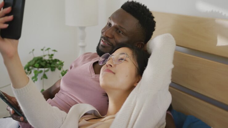 Happy diverse couple using smartphone and lying in bedroom