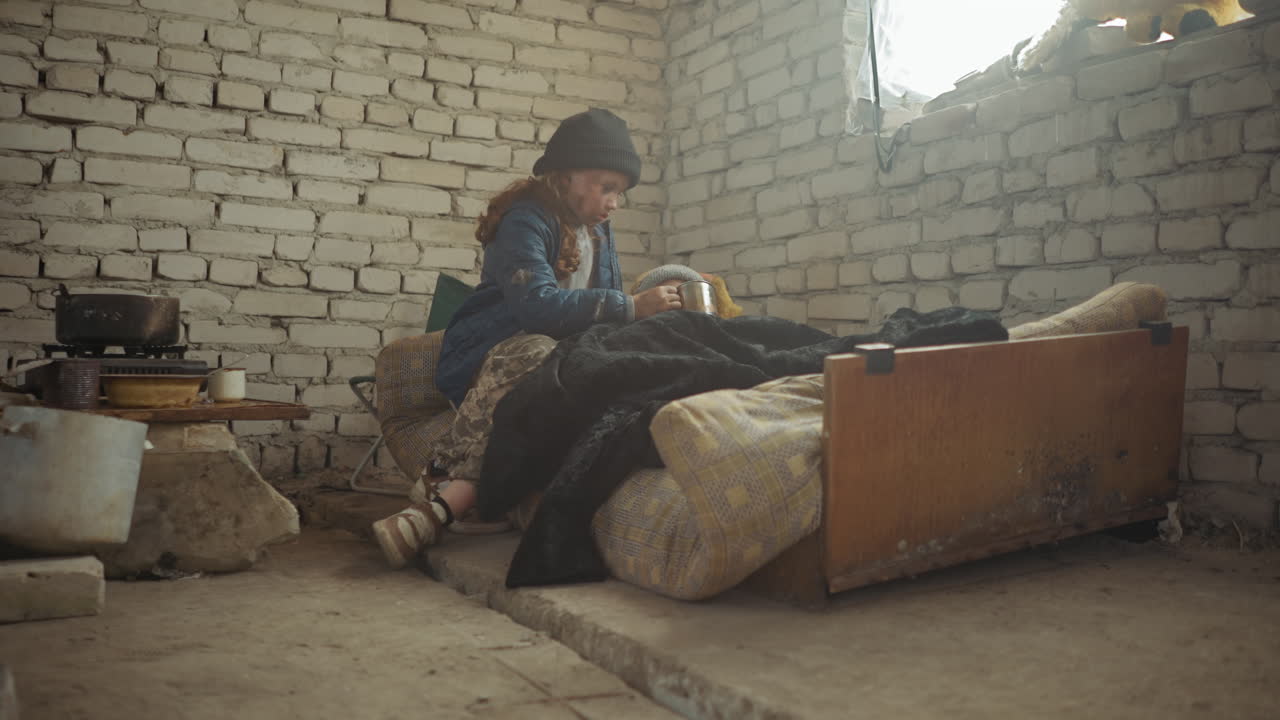 Dirty orphan girl in beanie and camouflage pants offers metal cup of water to sick friend lying under blanket on torn mattress in cold brick room with scattered pots and stove, showing care
