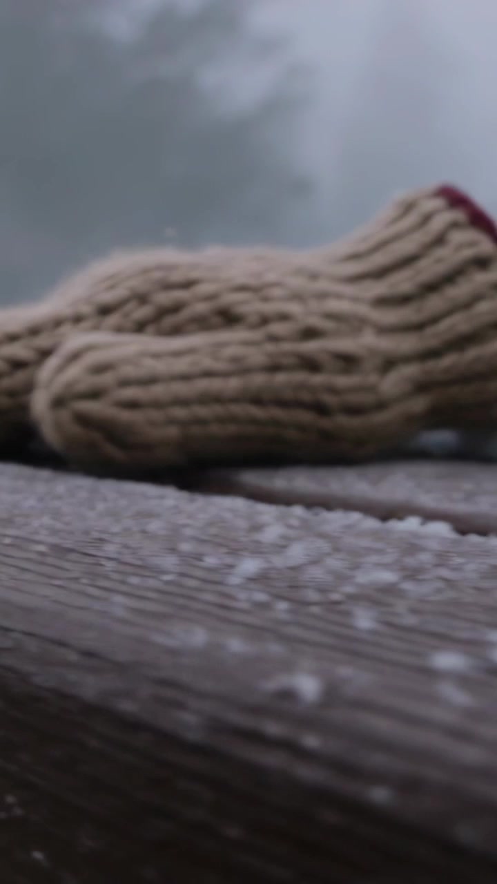 Video verticale: Entrando da destra, una mano con un guanto beige che spazza via la neve sottile su una ringhiera di legno.