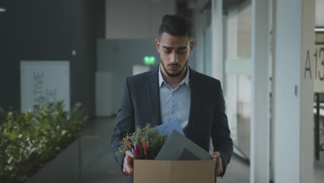 young upset middle eastern man manager being fired, walking at office building and carrying box with personal belongings