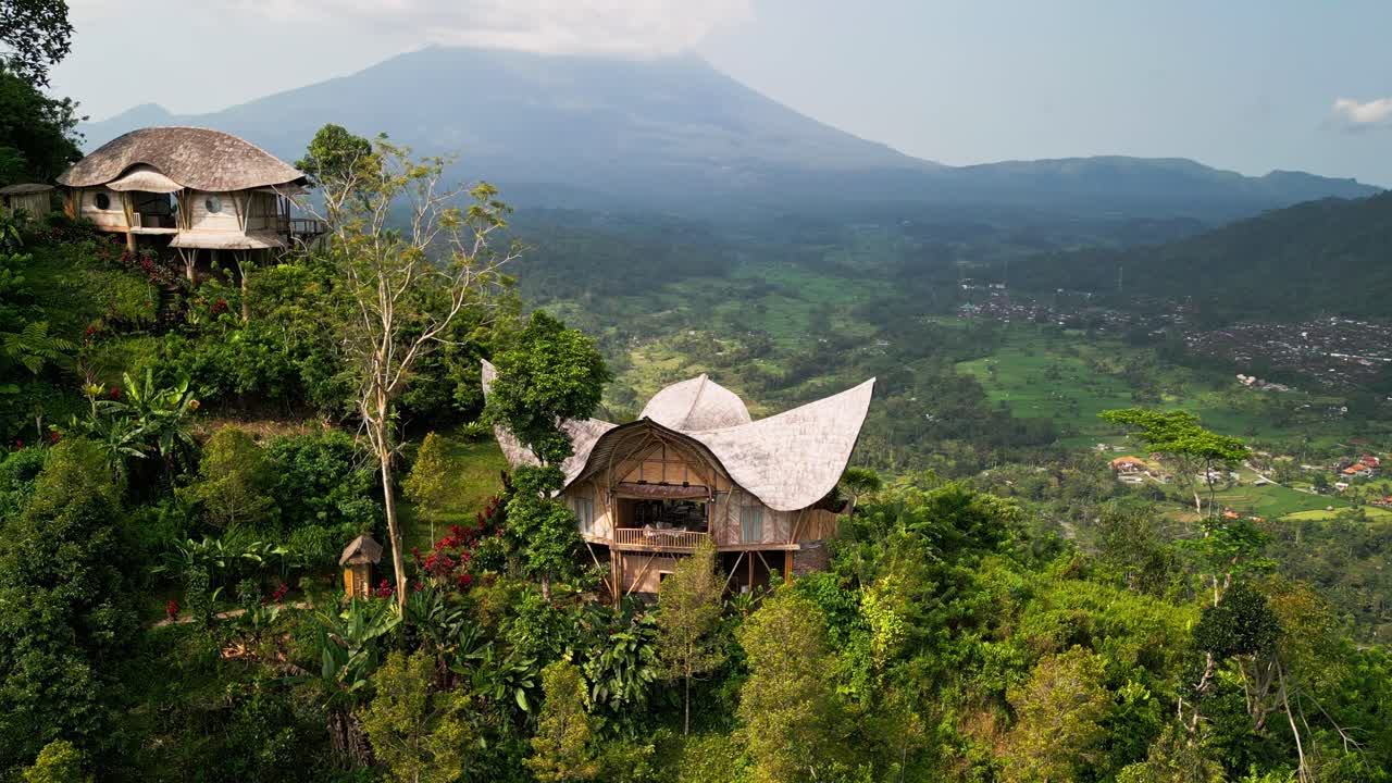 Drone footage of handcrafted bamboo villas perched on a lush Bali hillside, overlooking Mount Agung and endless green valleys, capturing the harmony of eco design and tropical nature