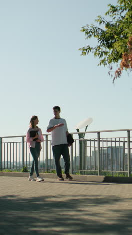 estudiantes caminando por un camino pavimentado cerca de una barandilla de hierro con libros en la mano, participando en una conversación alegre rodeados de vegetación, paisaje urbano y arquitectura urbana bajo la brillante luz del sol