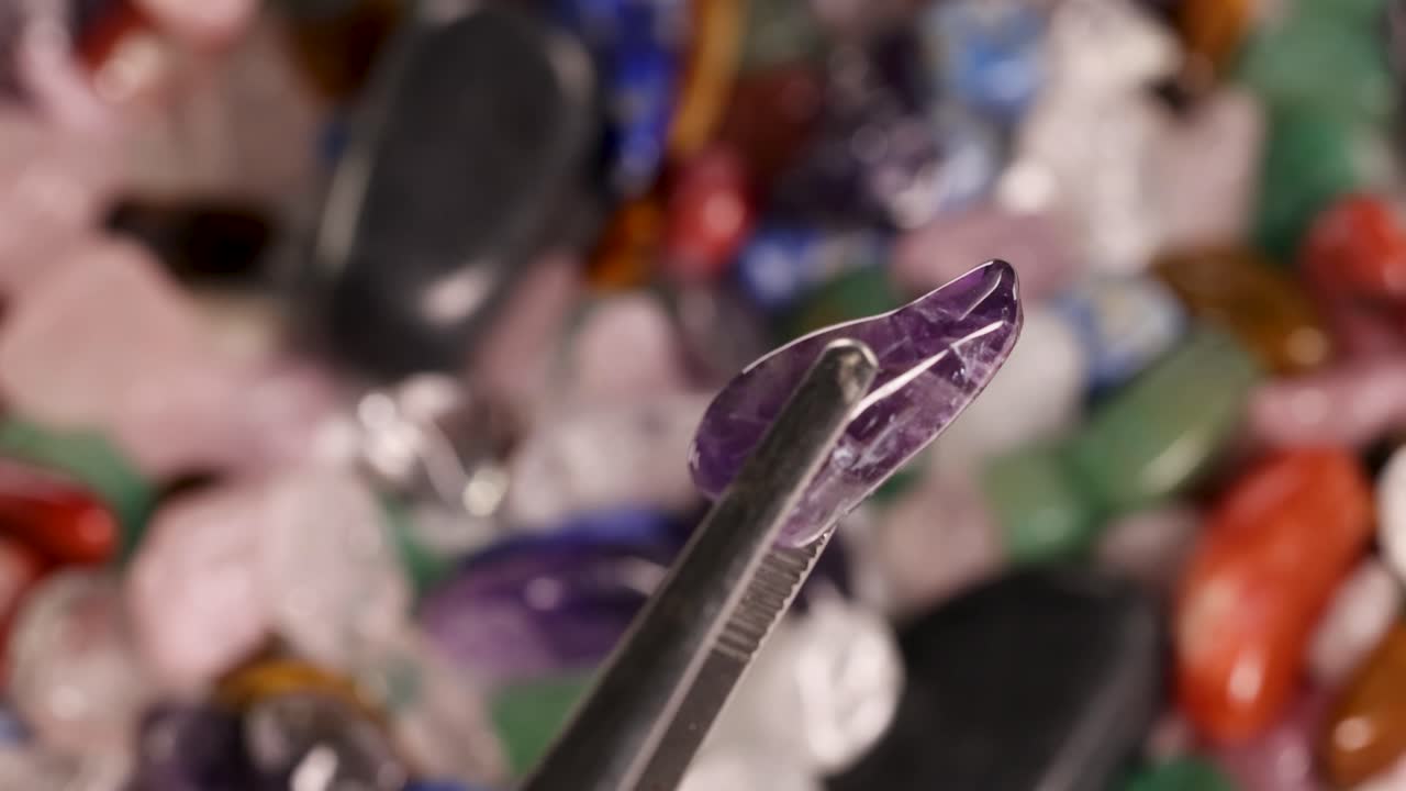 A close-up view of tweezers handling an amethyst stone amidst a colorful array of gemstones under warm lighting